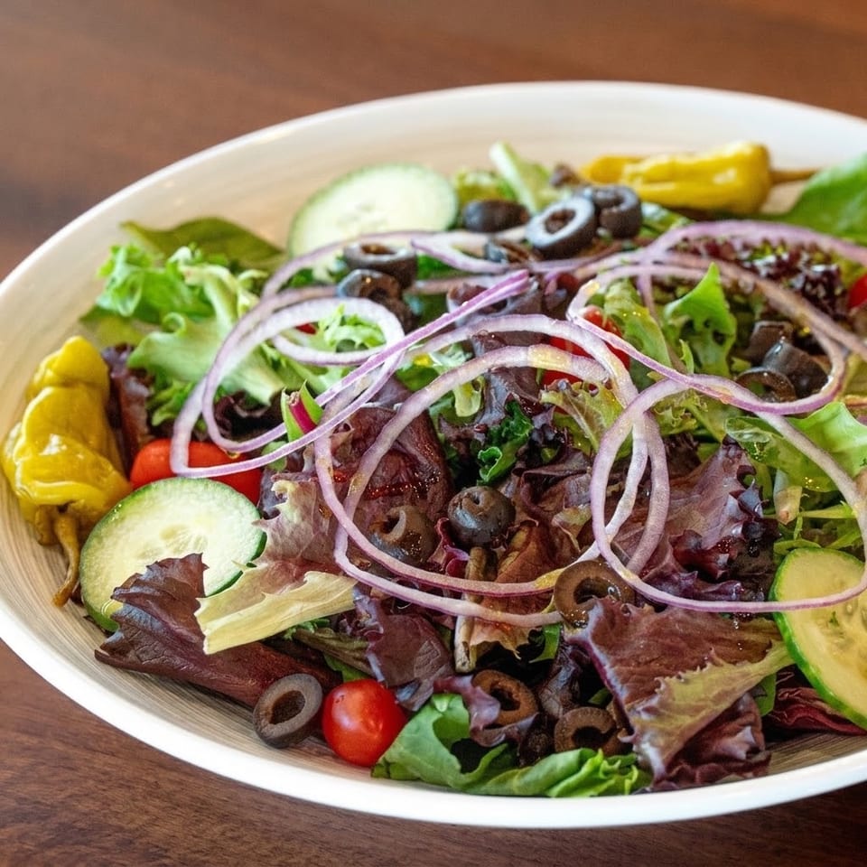 A fresh garden salad with mixed greens, red onions, olives, cucumbers, cherry tomatoes, and banana peppers.